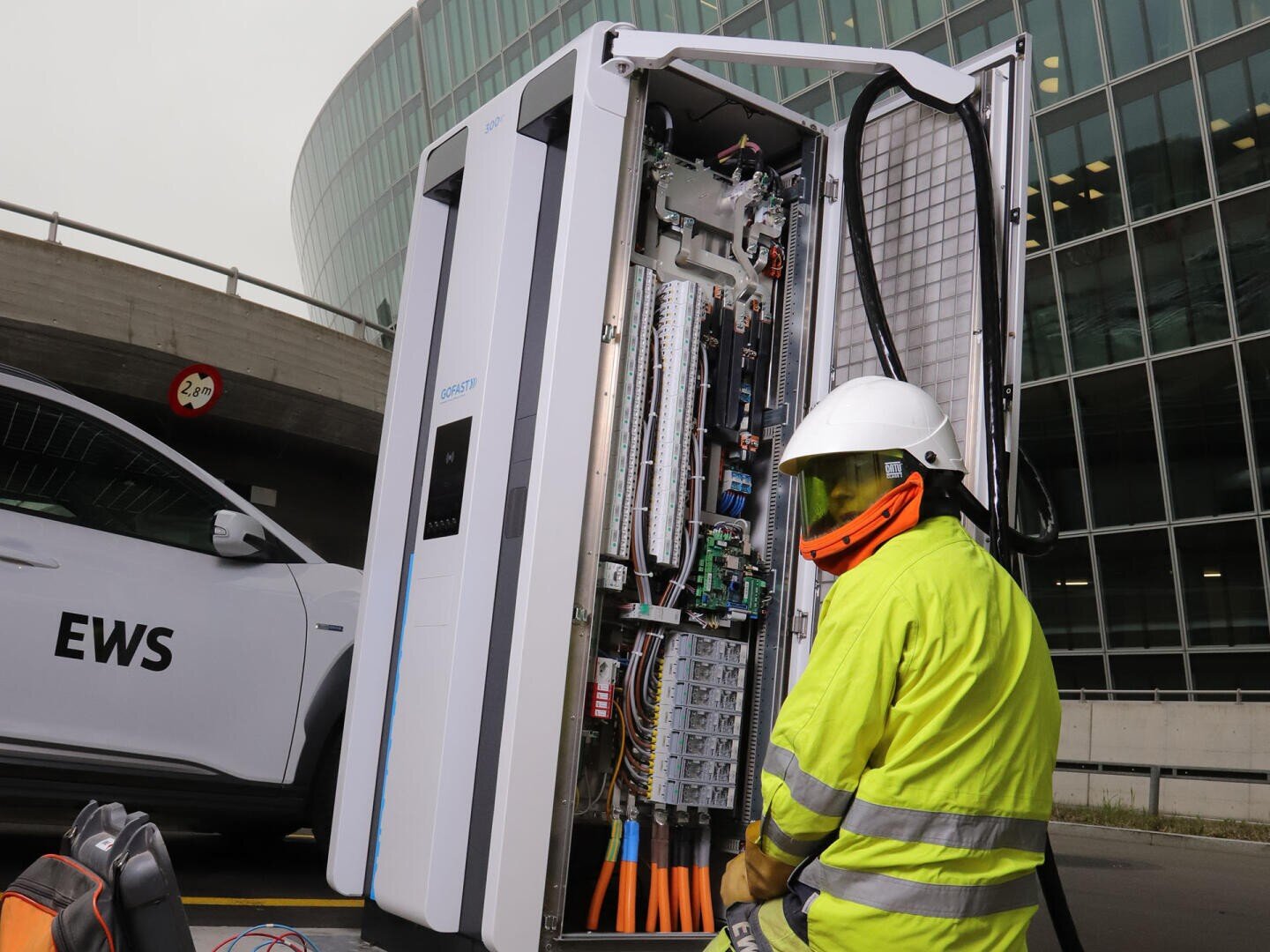 A worker in a high-visibility suit and helmet inspects an open switch cabinet outdoors. A building and a vehicle with EWS markings can be seen in the background.