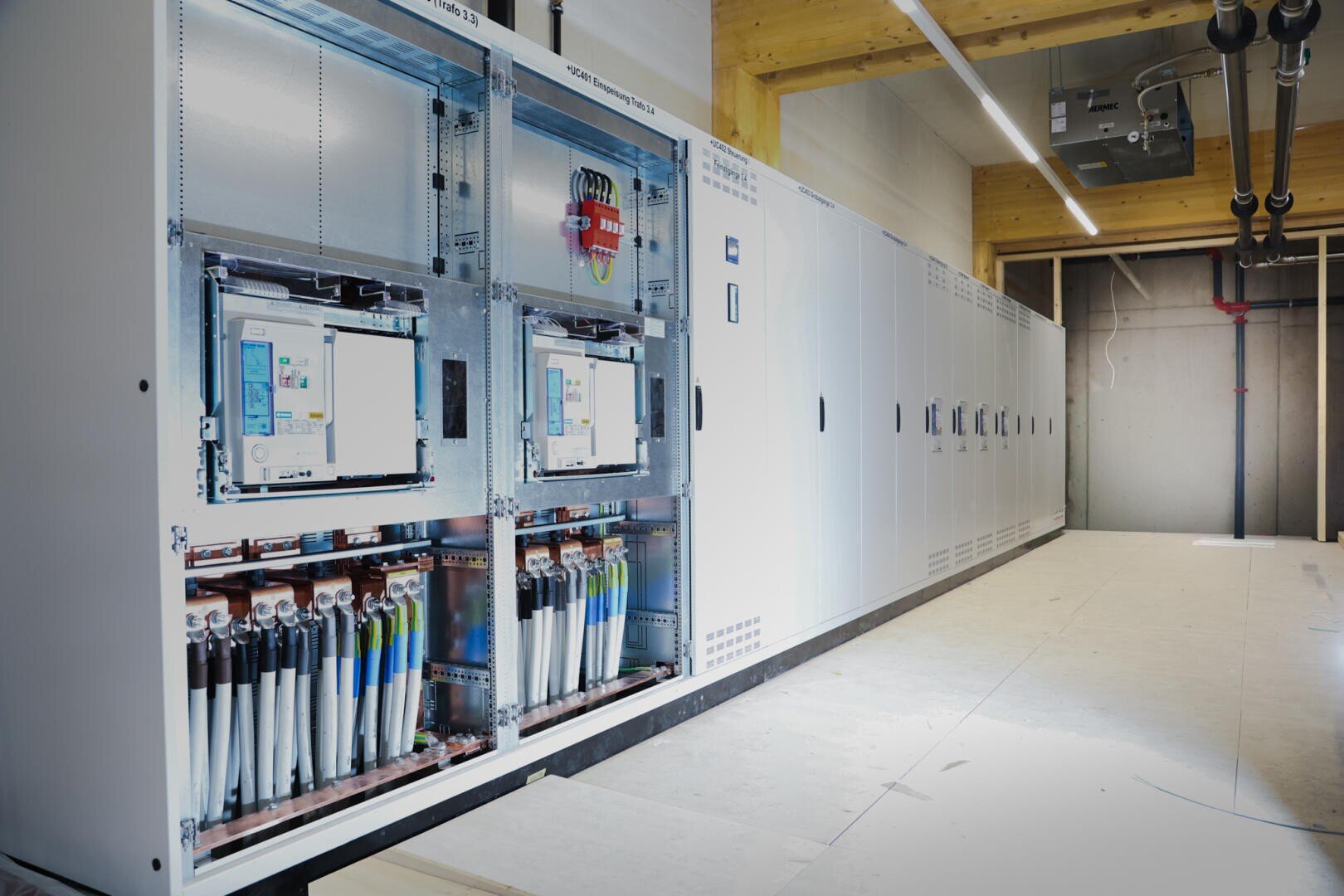 A brightly lit server or control room with rows of large white switch cabinets, some of which are open to reveal complex cabling, circuit boards and control panels inside. The floor is tiled and pipes are visible on the ceiling.