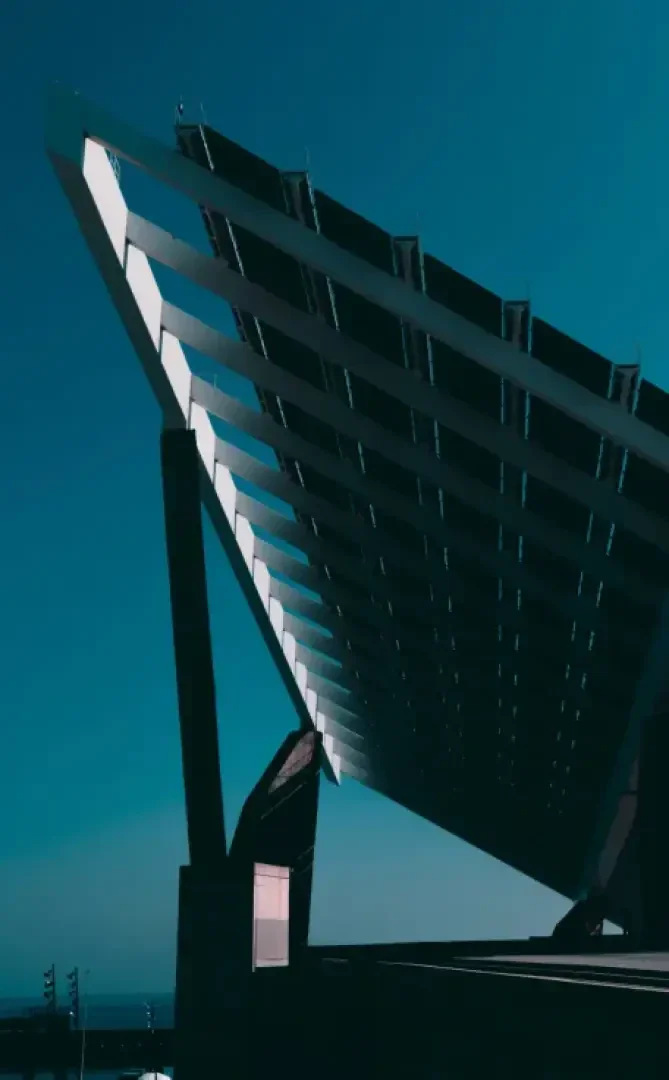 A large, modern solar panel array with angled, rectangular panels is elevated on sturdy supports against a clear blue sky, casting sharp geometric shadows below.