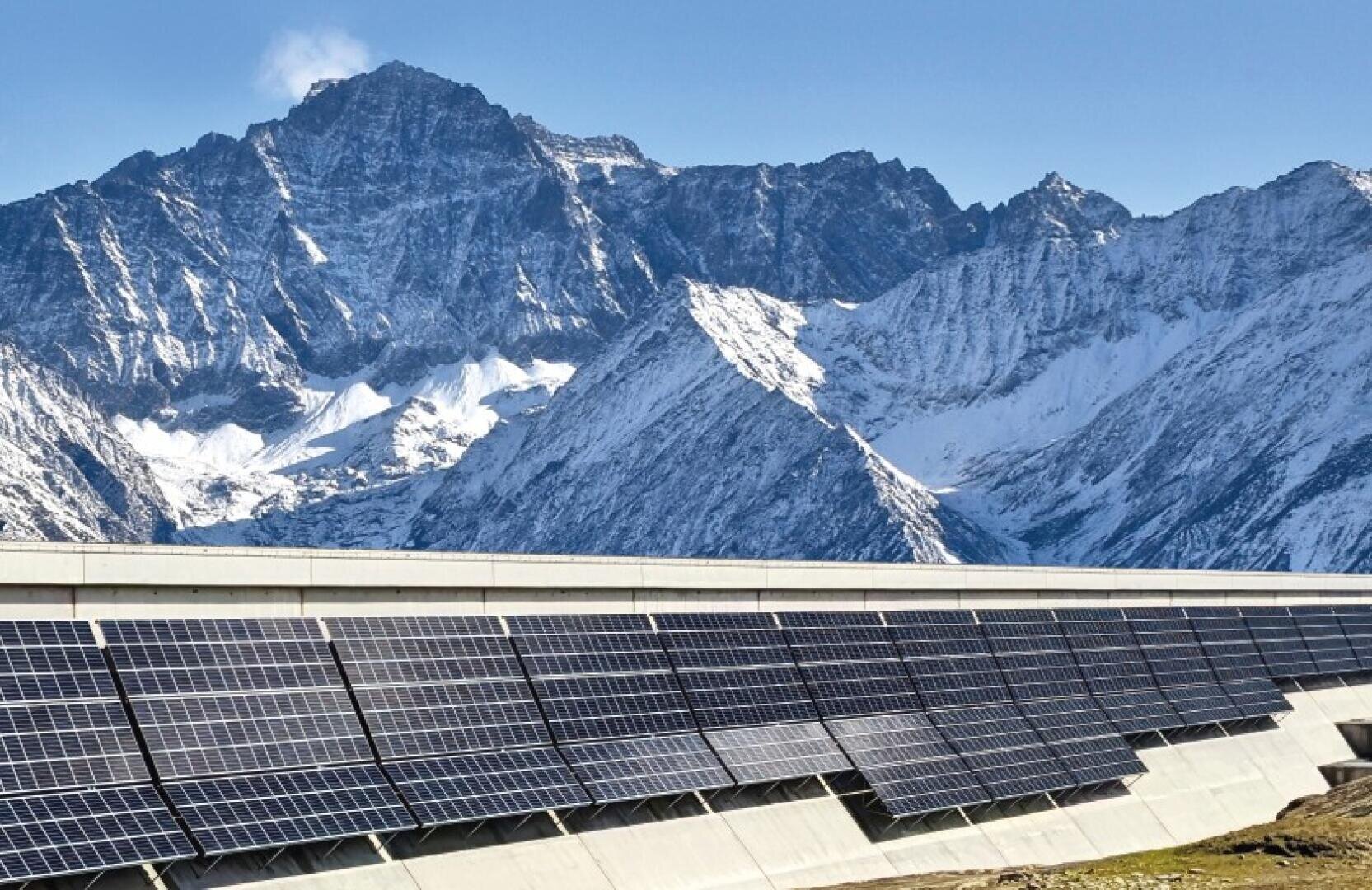 Reihen von Solarmodulen auf einer Betonplattform stehen vor der Kulisse hoher, schneebedeckter Berge unter einem strahlend blauen Himmel.