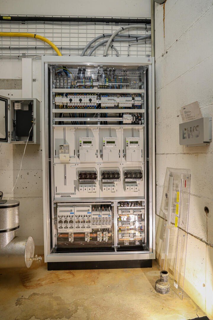 A large electrical panel with several switches, meters, cables and circuit breakers is mounted on a wall in an industrial plant, with pipes and cables running above and around it.
