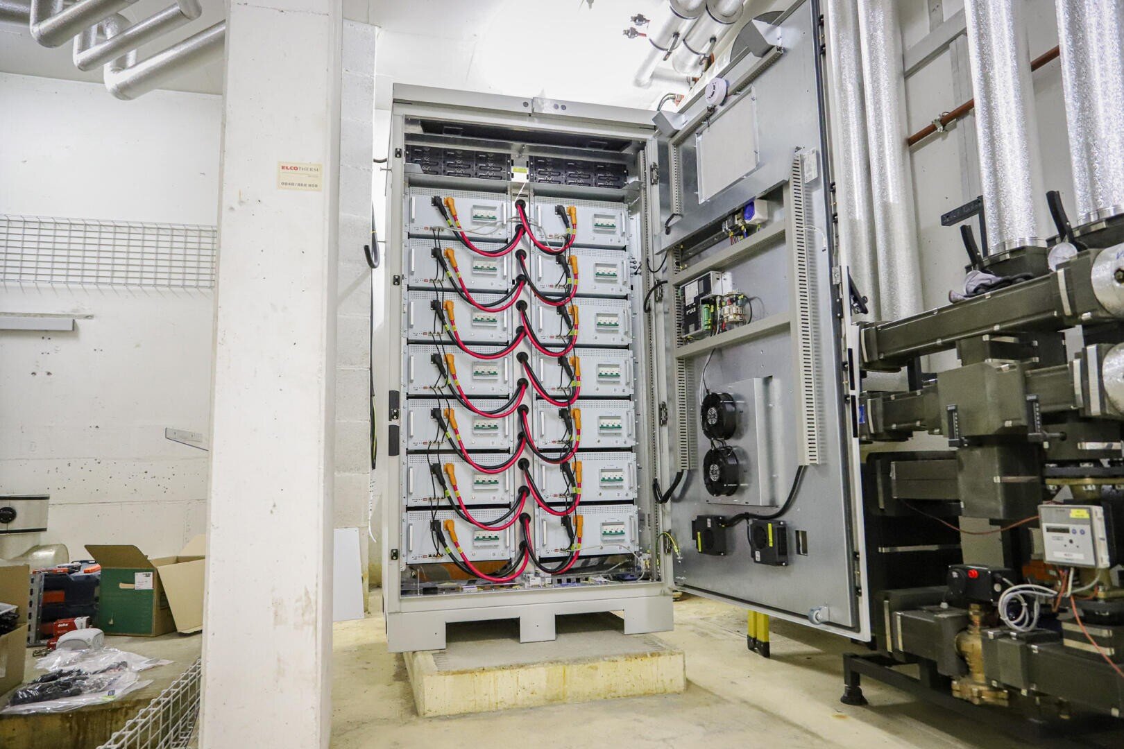 An open industrial electrical cabinet shows several stacked battery units connected with red and black cables, surrounded by pipes, machines and tools in a supply room.