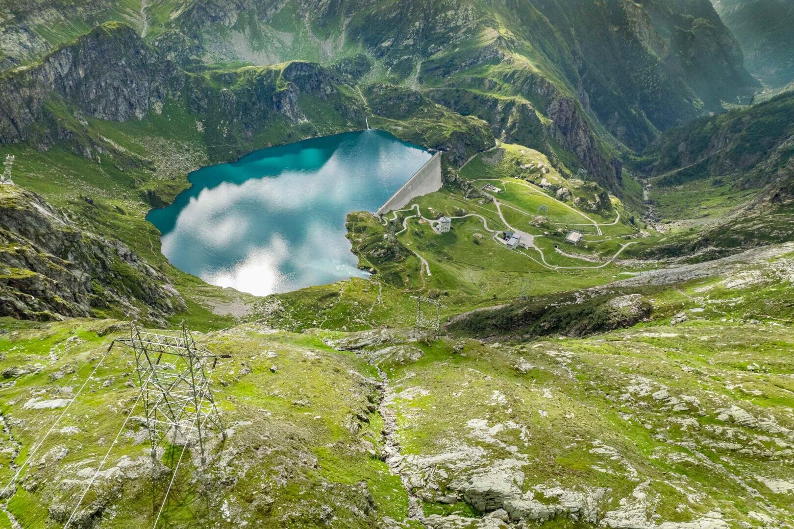Luftaufnahme eines türkisfarbenen Bergsees, umgeben von grünen Hügeln und felsigem Gelände, mit einem Staudamm, gewundenen Straßen, Stromleitungen und verstreuten Gebäuden unter einem teilweise bewölkten Himmel.