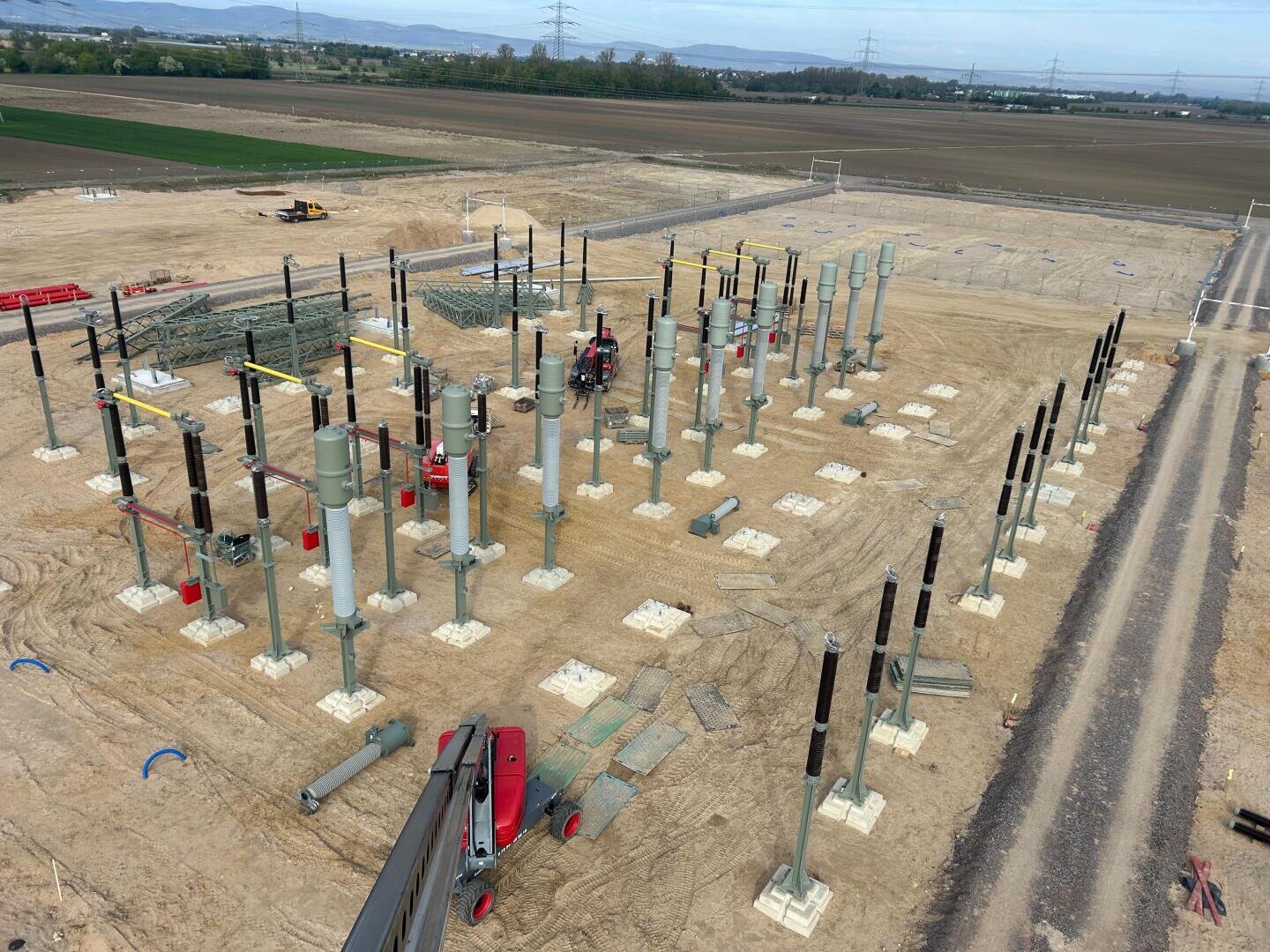 Aerial view of a substation under construction on an unpaved site with numerous steel pillars and insulators, surrounded by open fields and distant hills.