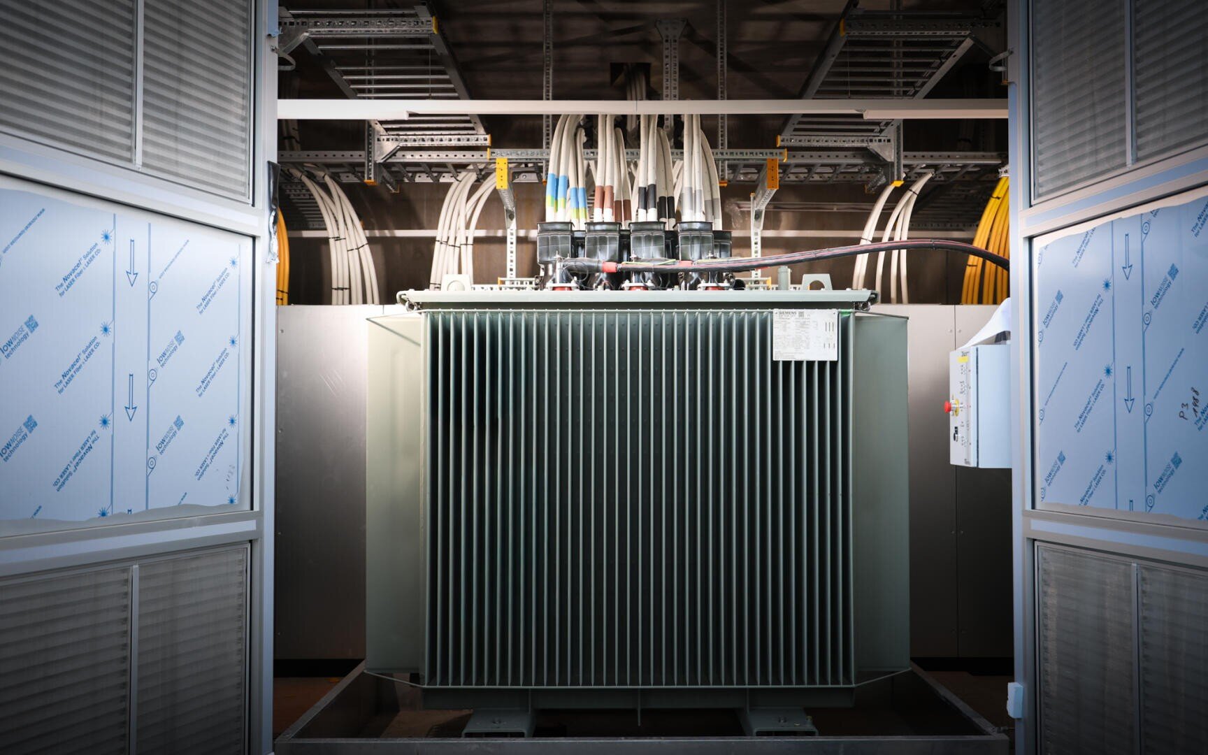 Large industrial electrical transformer with cooling fins, cables and wiring on top, in a brightly lit room with metal panels and visible conduits on the ceiling in the background.