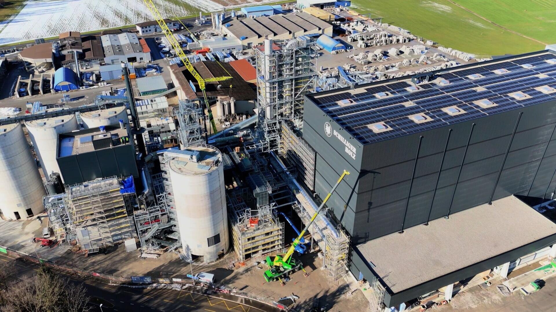 Aerial view of an industrial plant with large tanks, scaffolding and a black building with solar cells on the roof; construction vehicles and workers can be seen on the ground.