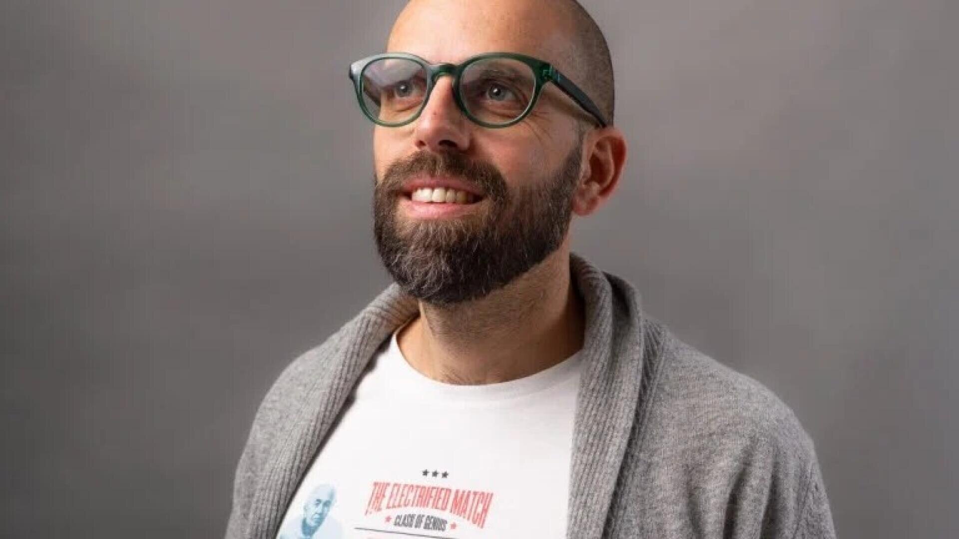 Ein bärtiger Mann mit Brille und rasiertem Kopf trägt eine graue Strickjacke über einem weißen bedruckten T-Shirt, lächelt und blickt leicht nach oben vor einem schlichten grauen Hintergrund.