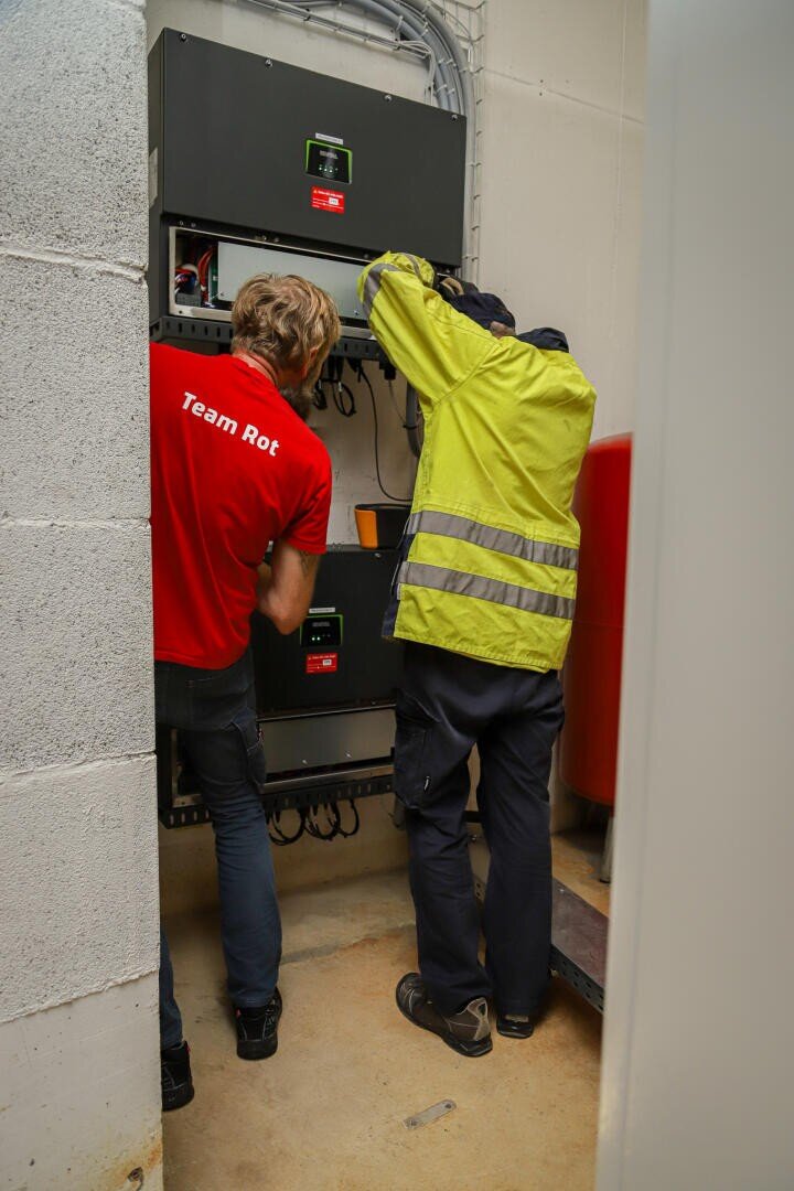 Two people are working on electrical appliances in a small utility room. One is wearing a red Team Red shirt, the other a yellow safety vest. Both are facing the appliances and concentrating on the wiring or the controls.