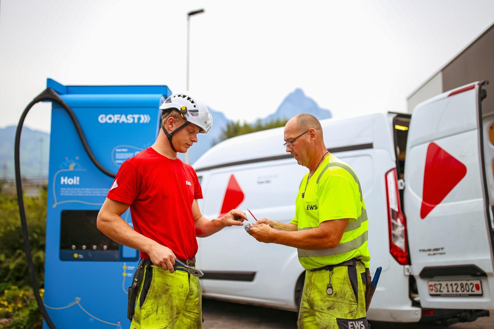 Two employees of an energy supplier, one in a red shirt and helmet, the other in a yellow shirt, stand next to a GOFAST charging station and white service trolleys and discuss paperwork or ID cards. The mountains can be seen in the background.
