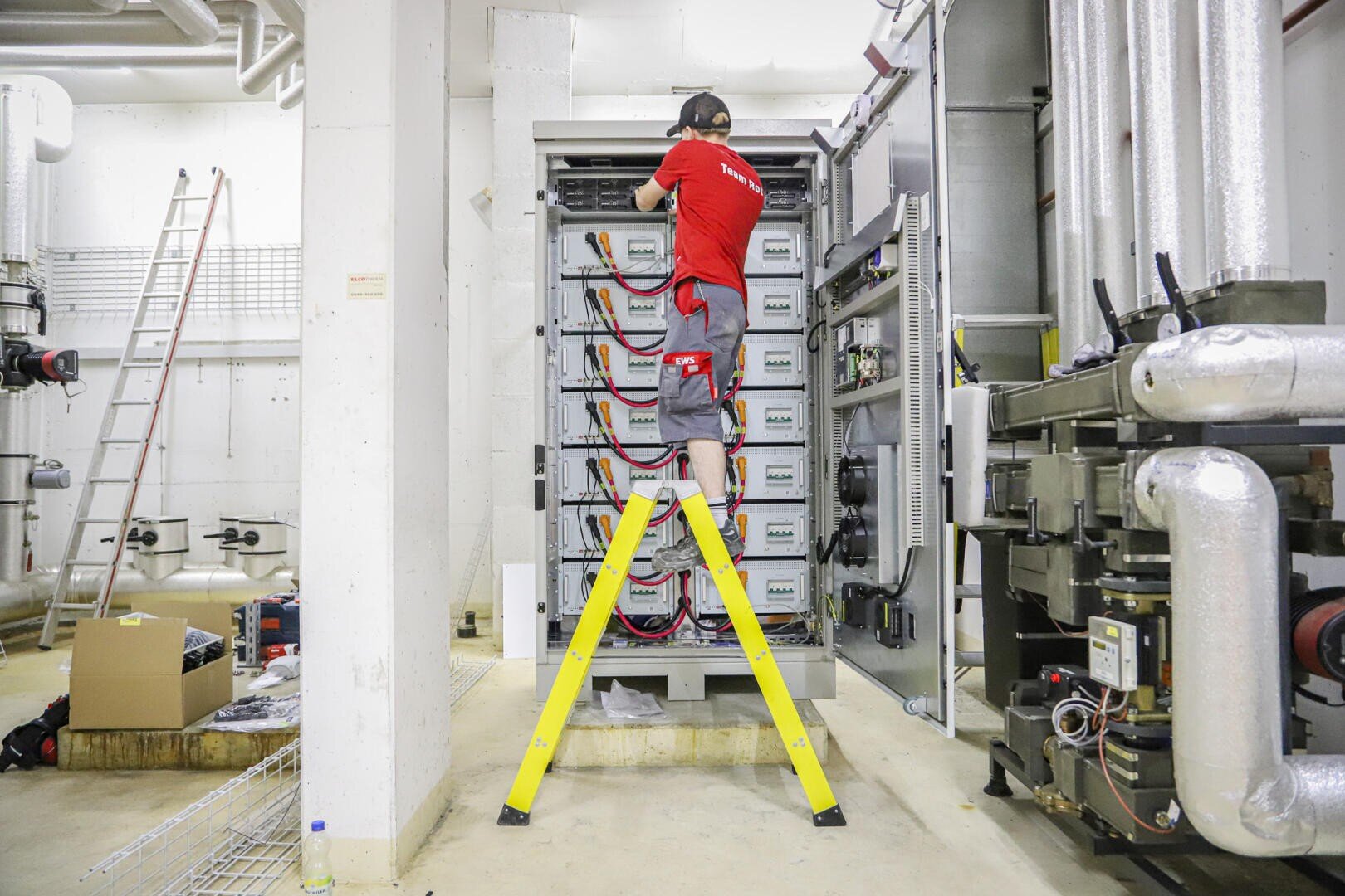 A technician in a red shirt stands on a yellow ladder and works on an electrical system in an industrial room with pipes and machines.