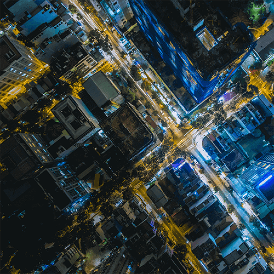 Aerial view of a city intersection at night, with illuminated streets forming an X shape and buildings lit by yellow and blue lights.