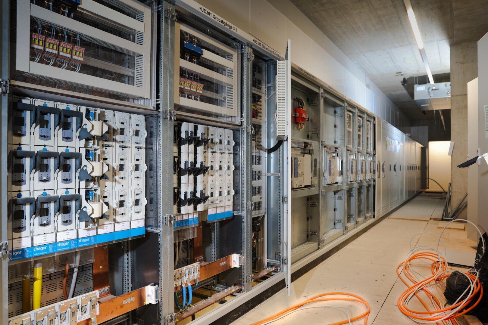 Rows of electrical panels and circuit breakers in an industrial space under construction with exposed wiring and orange cables on the floor. Overhead fluorescent tubes illuminate the unfinished space.