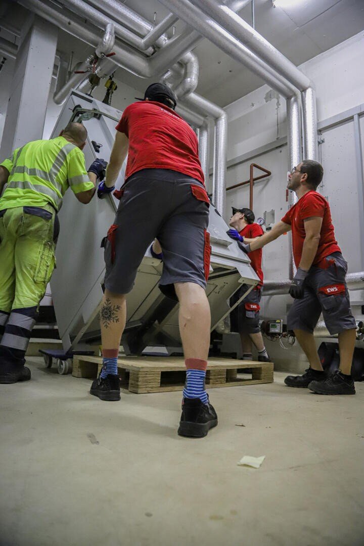 Four workers, three in red shirts and one in a yellow and green outfit, move a large, heavy machine on a pallet in an industrial space with exposed pipes and a concrete floor.