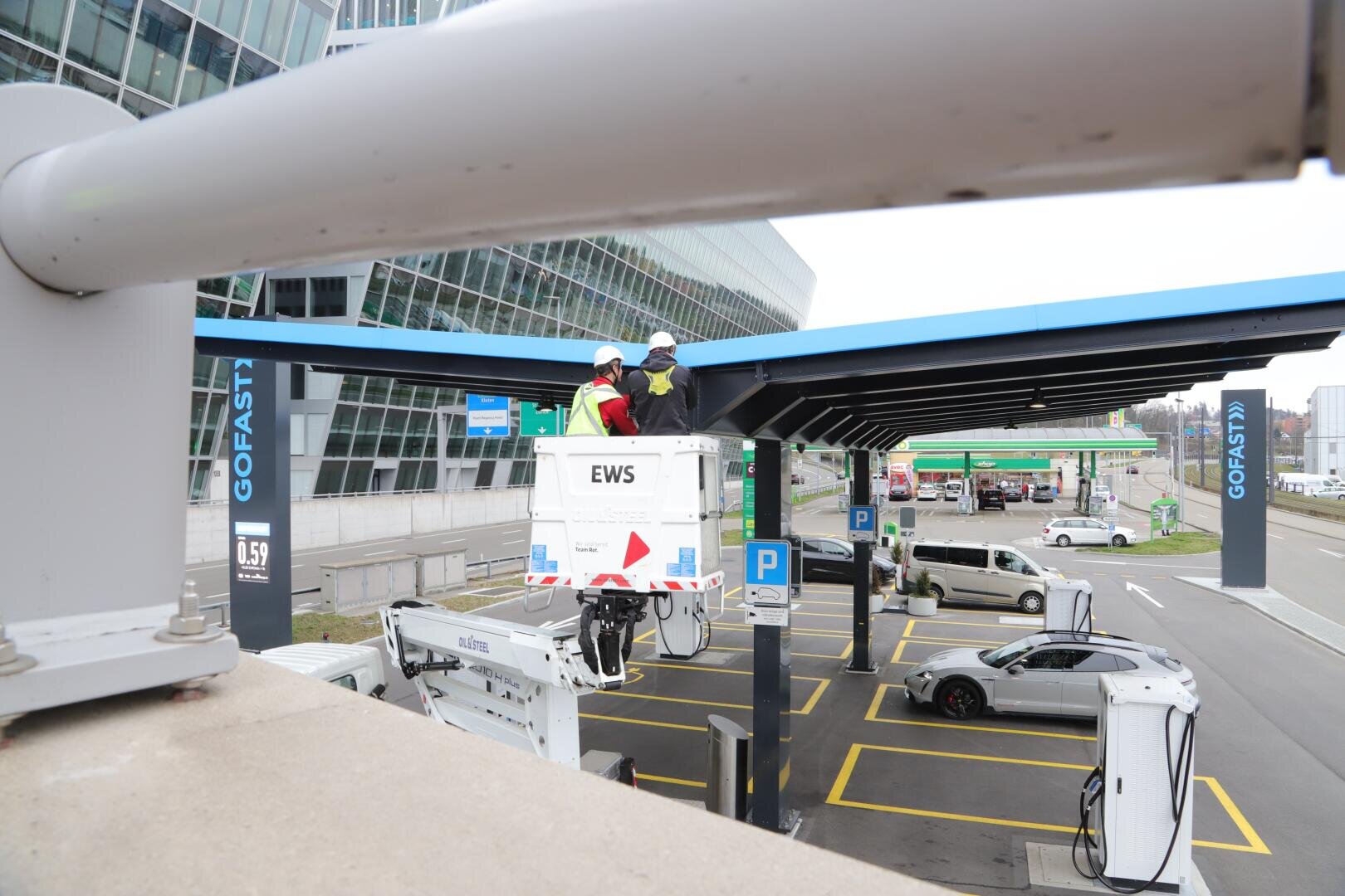 Two workers in an elevator are working on the canopy of a charging station for electric vehicles. Several white charging stations and cars can be seen below. The area is marked with yellow lines and signs and is located near a modern glass building.