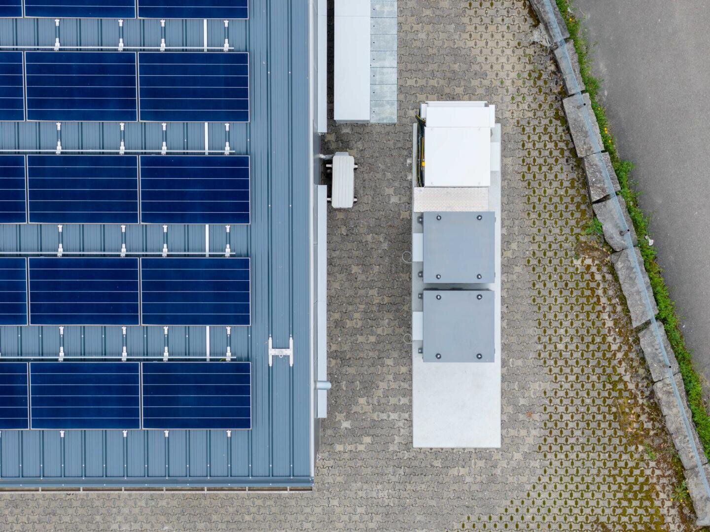 Luftaufnahme von blauen Solarmodulen auf einem Metalldach neben einer gepflasterten Fläche mit Stromkästen und einer von Betonbordsteinen begrenzten Fahrbahn.