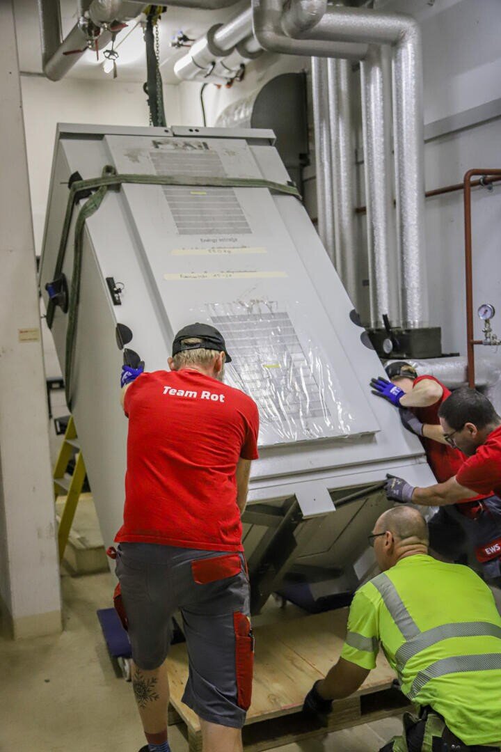 Three workers in protective clothing guide a large, tilted industrial machine onto a wooden platform in an engine room with exposed pipes. Two are wearing red shirts labeled "Team Red" and one is wearing a high visibility yellow shirt.