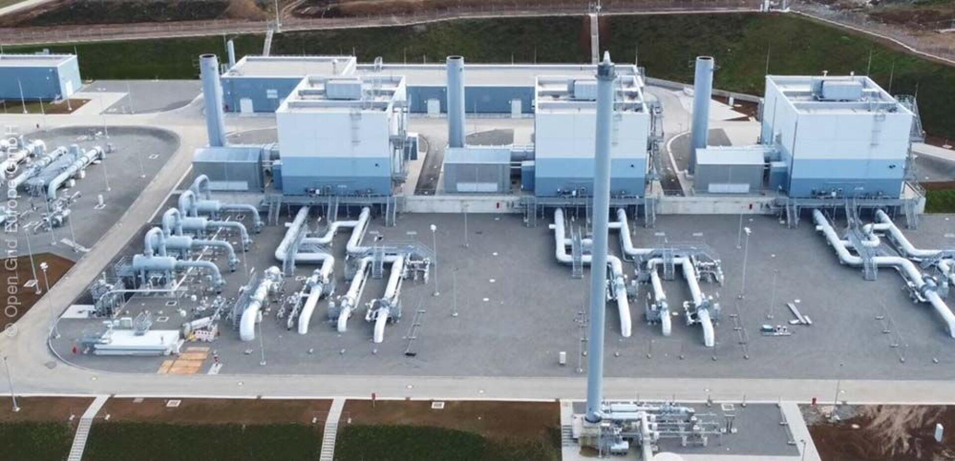 Luftaufnahme einer großen Industrieanlage mit mehreren Gebäuden, Schornsteinen und einem ausgedehnten Netz von großen Rohrleitungen, die in geordneten Reihen auf einer gepflasterten Fläche angeordnet sind. Grünes Gras umgibt den Komplex.