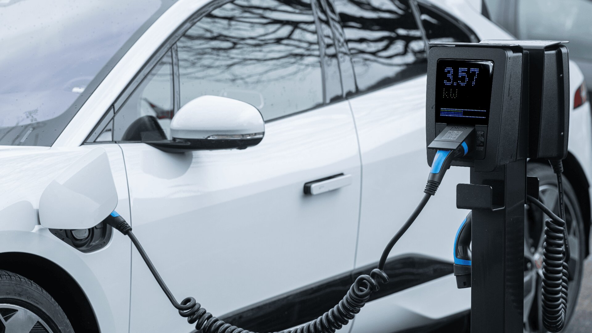 Ein weißes Elektroauto ist geparkt und an eine Ladestation angeschlossen, wobei der Bildschirm des Ladegeräts Informationen zum Ladevorgang anzeigt. Baumzweige spiegeln sich auf dem Autofenster.