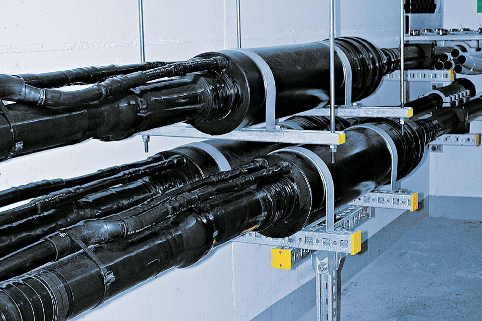 Zwei große schwarze Kabel sind mit Metallbügeln sicher an einer Wand und am Boden in einer Industrieumgebung befestigt, wobei mehrere kleinere Kabel und Stecker entlang ihrer Länge sichtbar sind.