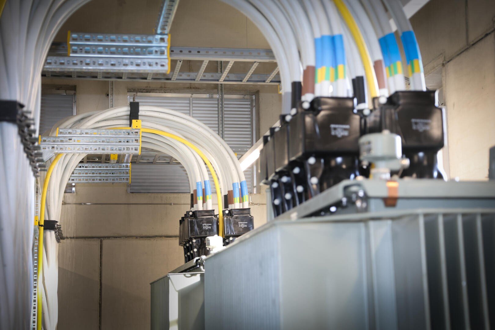 Large electrical cables and connectors are attached to industrial transformers in a utility room, with cable trays overhead and concrete walls in the background.