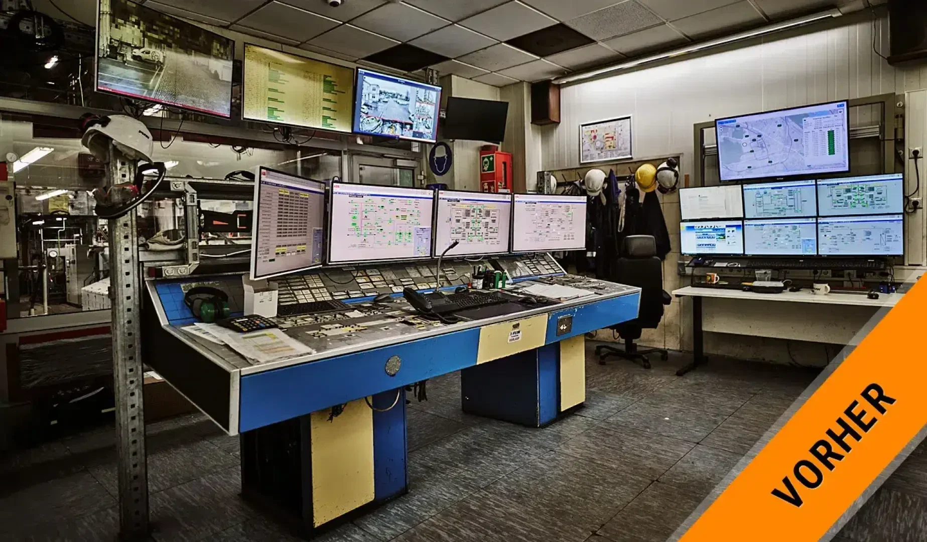 A control room with several monitors displaying data and surveillance material, various devices on desks, hard hats on shelves and an orange VORHER banner in the corner.