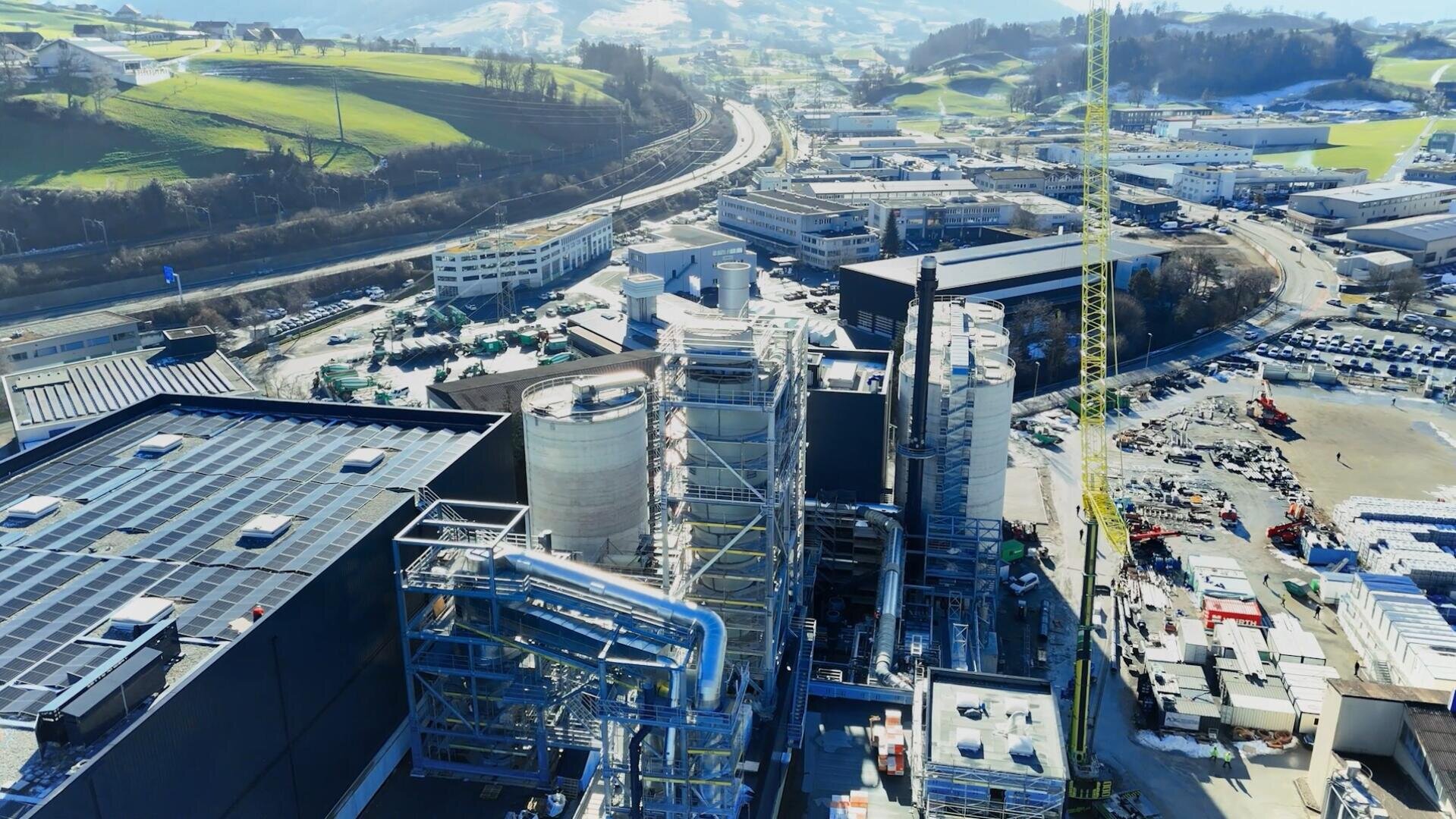 Aerial view of an industrial plant with large tanks, pipes and structures, surrounded by roads, buildings, parked vehicles and green hills under a clear sky.