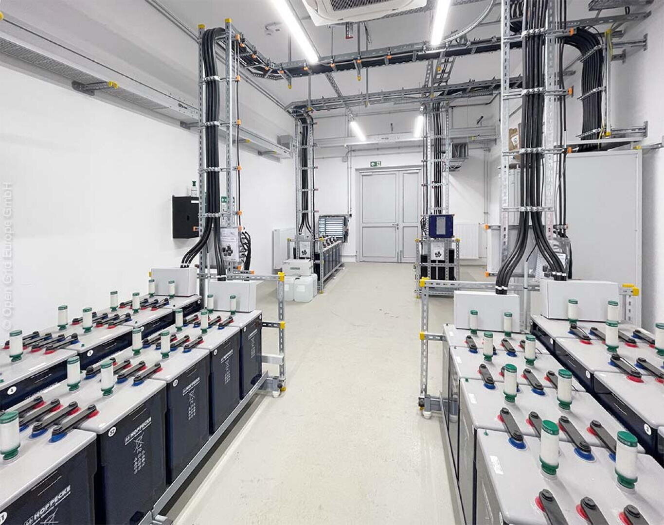 Ein sauberer, weißer Raum mit großen Batterieeinheiten, die durch Kabel und Metallrohre an der Decke und an den Wänden miteinander verbunden sind; im Hintergrund sind Leuchtstoffröhren und weiße Türen zu sehen.