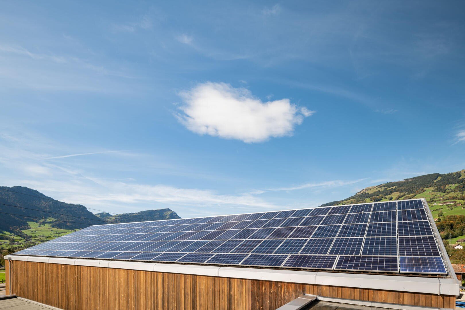 Auf dem Dach eines Holzhauses installierte Solarmodule mit Bergen, grünen Hügeln und einem blauen Himmel mit einer einzelnen weißen Wolke im Hintergrund.