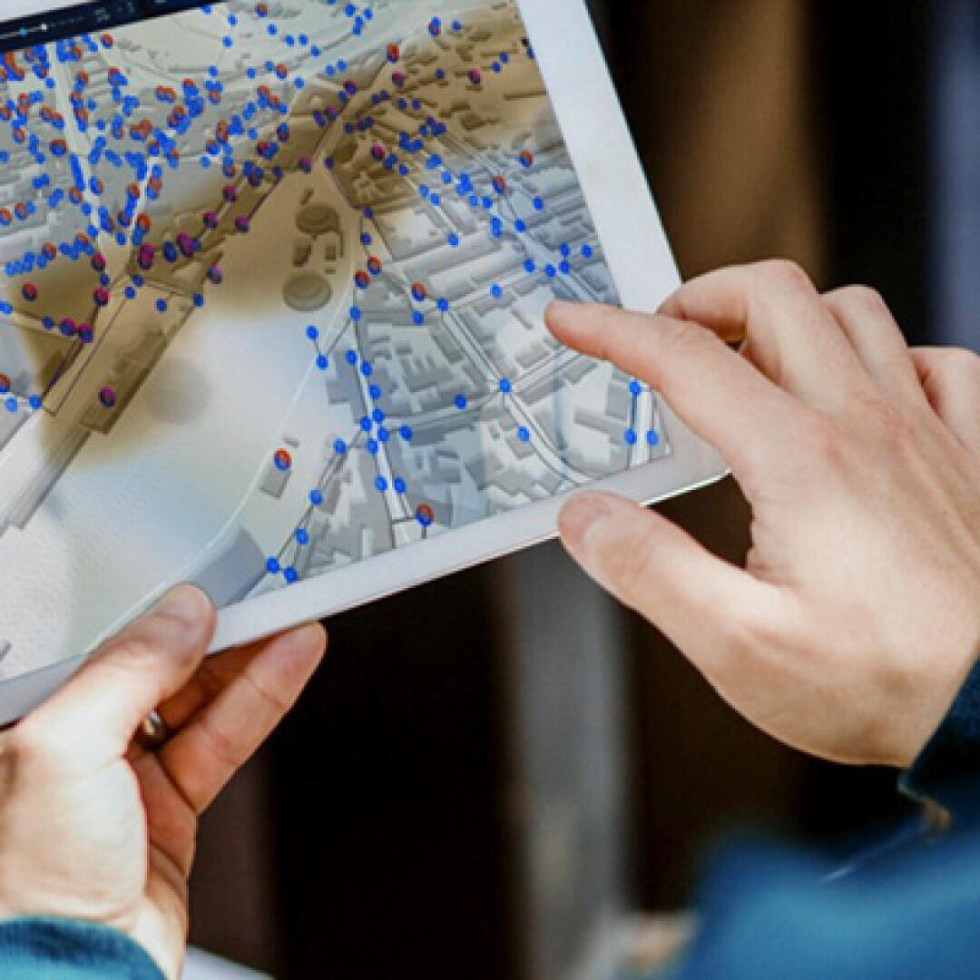 Eine Person benutzt ein Tablet, auf dem eine digitale Karte mit zahlreichen blauen und roten Datenpunkten und einem 3D-Layout von Gebäuden angezeigt wird.