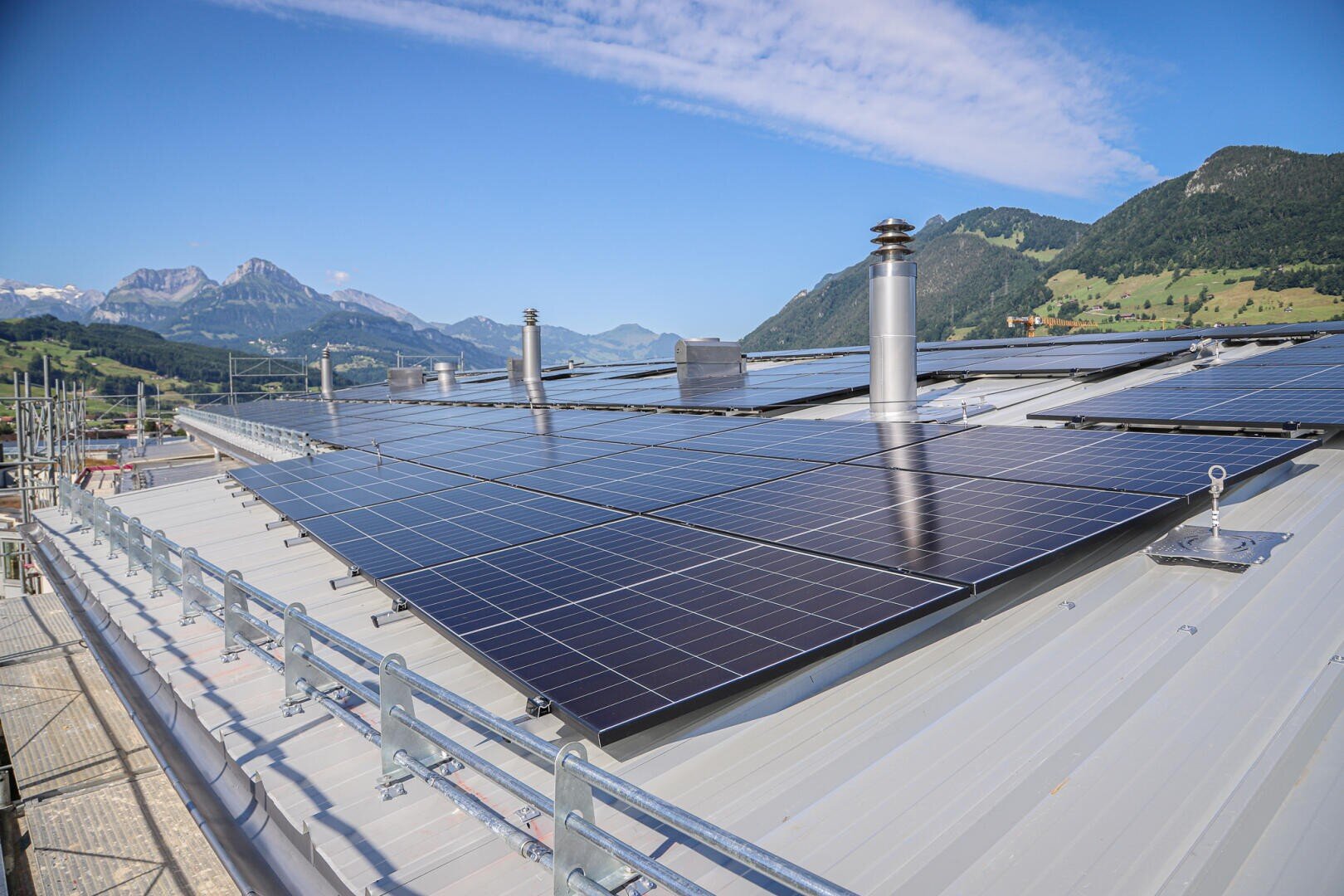 Rows of solar panels installed on a metal roof with chimneys and mountains in the background under a bright blue sky with scattered clouds.