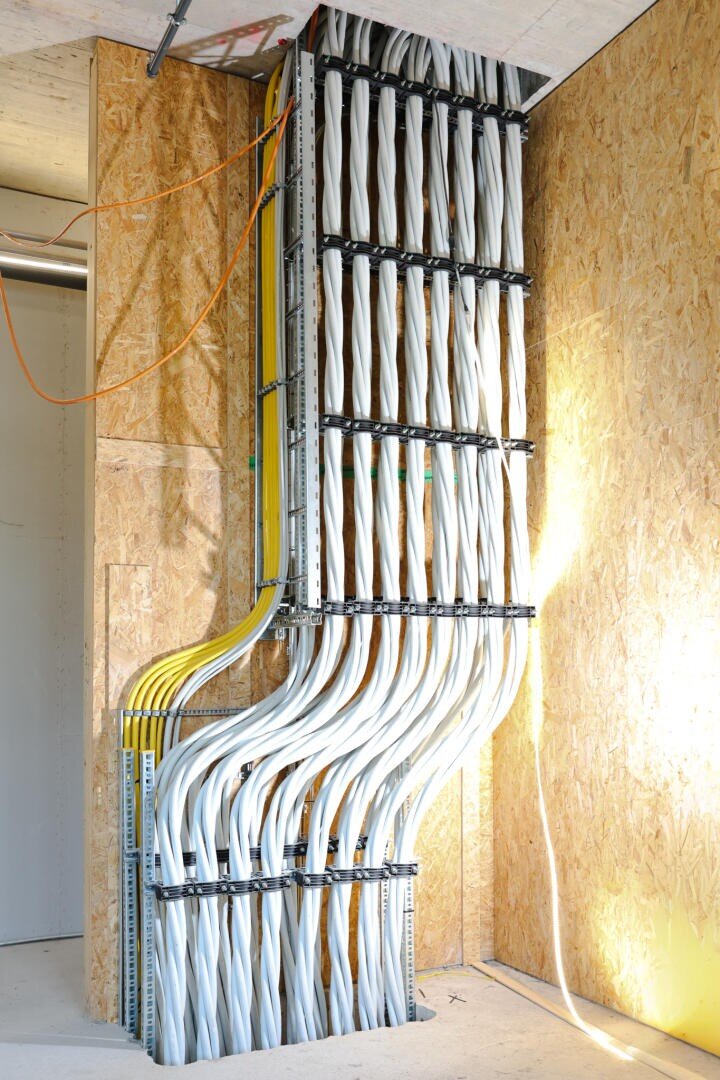 A neatly arranged bundle of white and yellow electrical cables runs vertically up a wall and is fastened with metal clips in a modern building with exposed walls made of stranded planks.