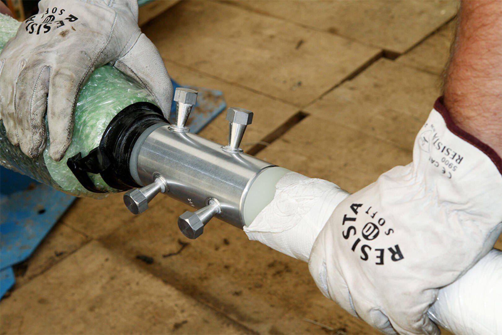 Zwei behandschuhte Hände verbinden zwei Rohre mit einer Metallkupplung mit Bolzen auf einer Holzfläche. Die Handschuhe sind mit Resista Soft beschriftet.