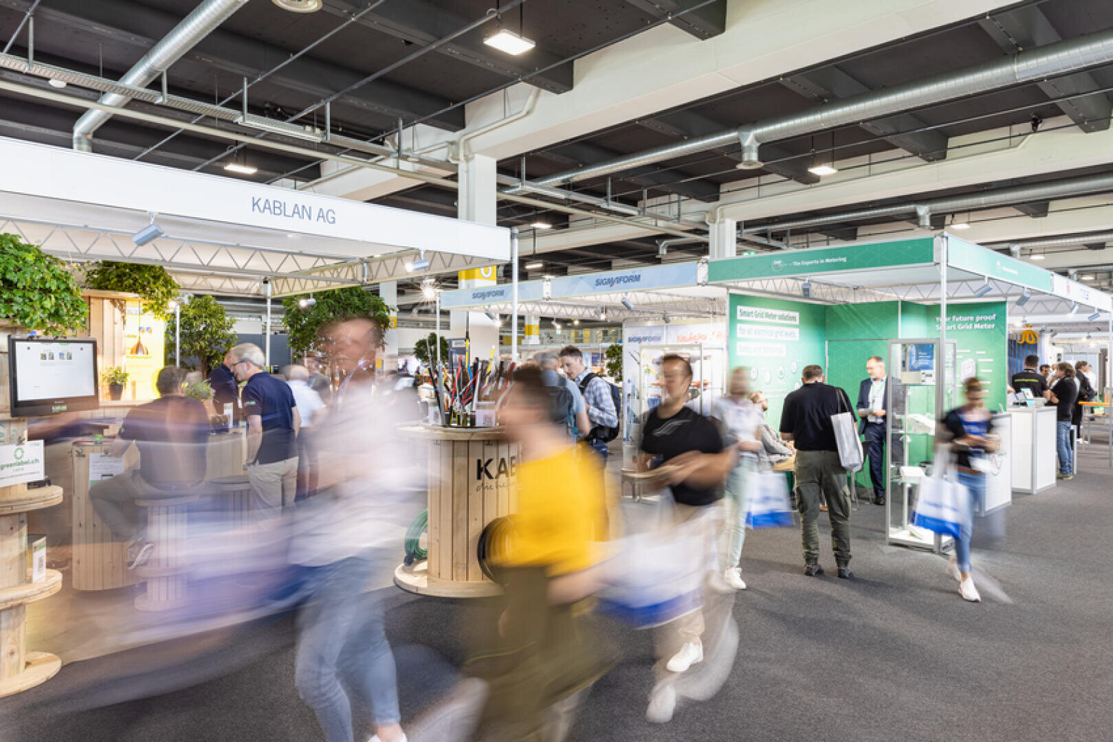 A busy indoor trade show with people walking past various exhibition booths, including one for KABLAN AG. Some visitors are blurred in motion, carrying bags, while others interact at the booths. The atmosphere is lively and bustling.