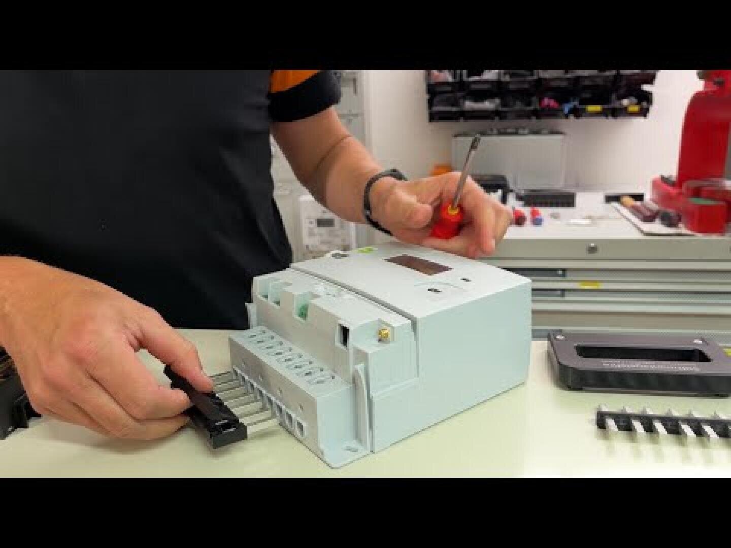 Eine Person benutzt einen Schraubendreher, um ein elektrisches Bauteil oder einen Schutzschalter auf einer Werkbank zu montieren oder zu reparieren, wobei verschiedene Werkzeuge und Teile im Hintergrund zu sehen sind.