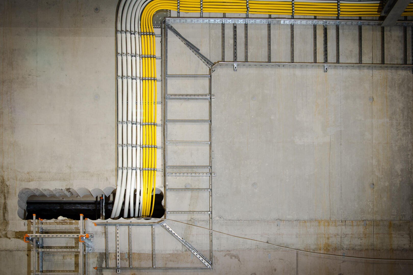 A series of yellow and white cables, neatly arranged on metal cable trays, run along a concrete wall and ceiling, making sharp right-angled bends. The cable trays are held in place by metal brackets.