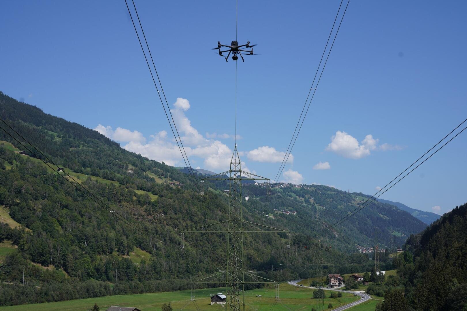 A drone hovers over power lines and a transmission tower in a green mountain valley, with a winding road and scattered houses under a clear blue sky.