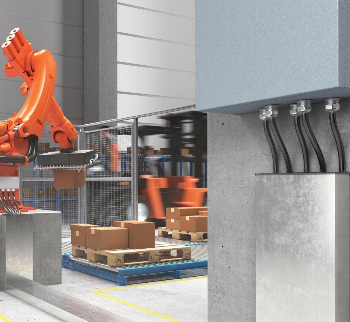 A robotic arm moves brown cardboard boxes on wooden pallets in a factory. The scene shows metal fences, industrial equipment and electrical cables attached to a wall-mounted switch box.