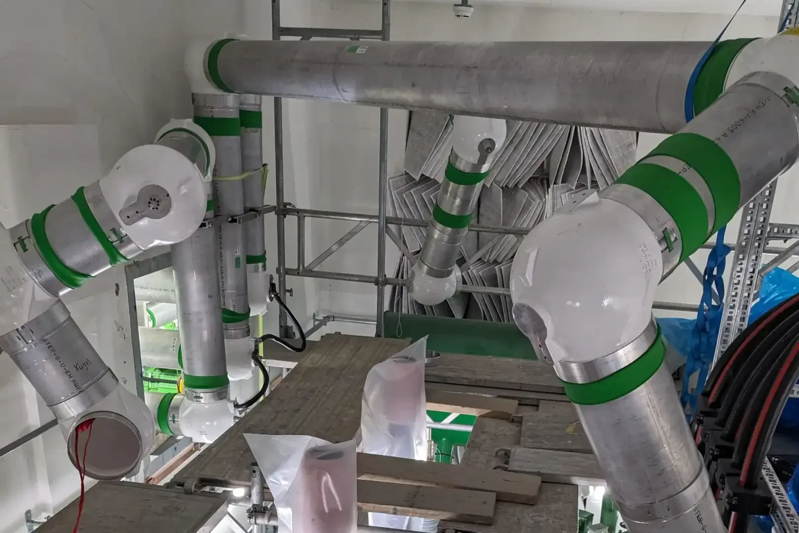 Industrial pipes with white and green markings run through a construction site surrounded by scaffolding and platforms. Some pipes have angled sockets, and sacks or containers are placed on wooden planks underneath.
