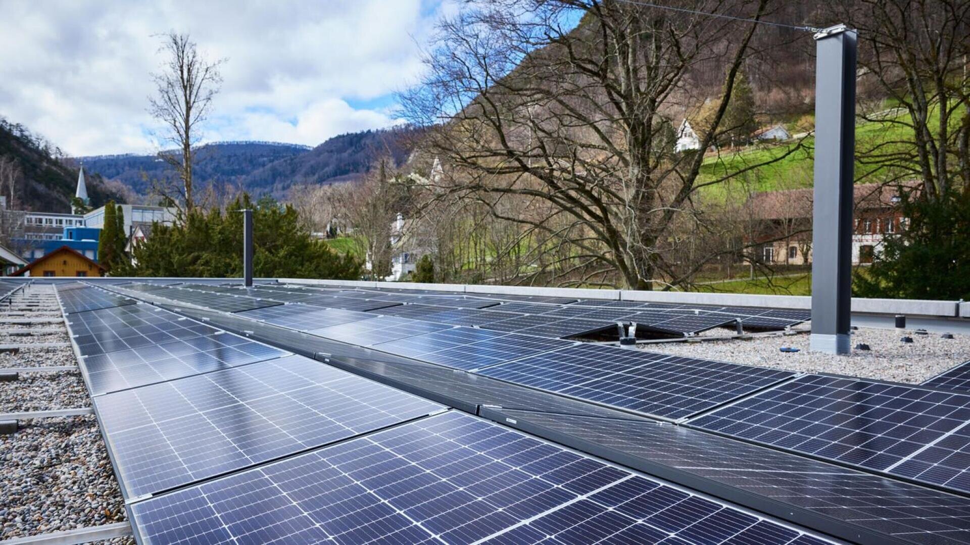 Rows of solar panels installed on a flat, gravel rooftop with trees and green hills in the background under a partly cloudy sky.