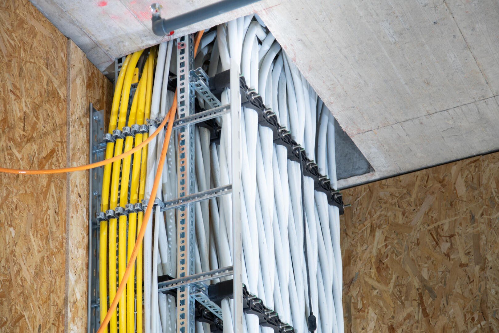 Several orderly bundles of yellow, white and orange power cables run vertically along a metal cable tray in a building under construction, surrounded by wooden and concrete walls.