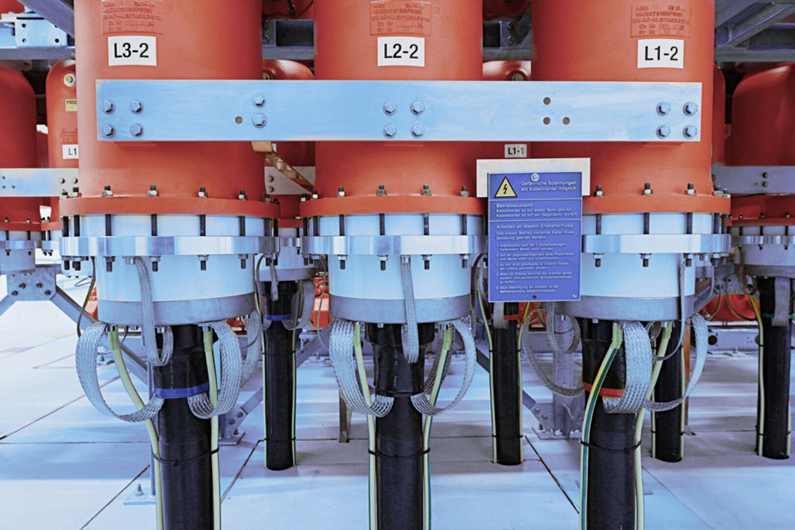 Nahaufnahme einer industriellen elektrischen Anlage mit orangefarbenen zylindrischen Isolatoren mit den Bezeichnungen L3-2, L2-2 und L1-2, die durch dicke Kabel und Metallstützen miteinander verbunden sind, und einem Warnschild, das an der mittleren Komponente angebracht ist.