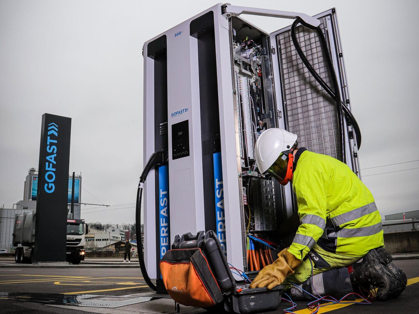 A worker wearing a yellow protective jacket and helmet repairs an open charging station for electric vehicles with tools and cables on the ground, near a GOFAST sign in an industrial area.